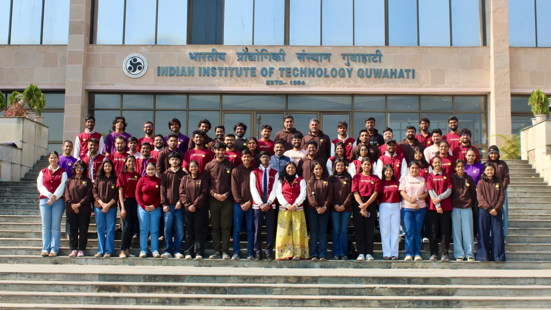 SPIC Macay IIT Guwahati team group photo on the steps of the main building in maroon oversized tees and coffee-brown hoodies