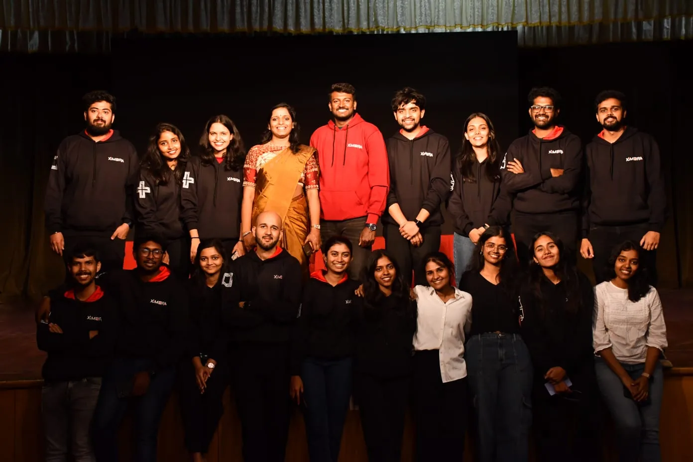 Ximera 2025 XIME Bengaluru core team group photo with 50+ members in matching black and maroon personalised hoodies on auditorium stage